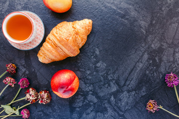 Breakfast with fresh croissant, cup of tea, peach and flowers on black background. Food background with copyspace.