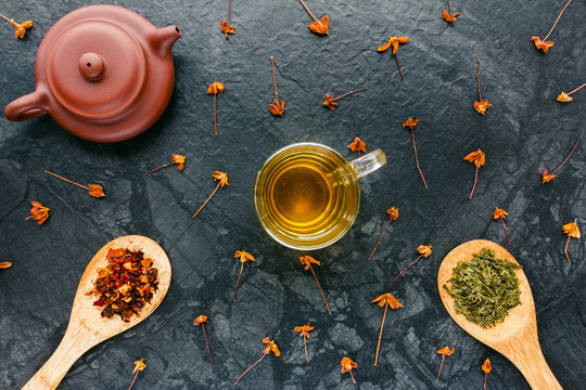 Herbal tea in glass, dried tea in wood spoons, teapot on black background