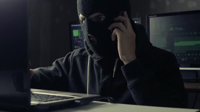 Dangerous hacker in a mask speaks phone at a computer in a dark office room