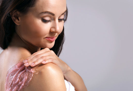 Young woman applying scrub on shoulder on grey background