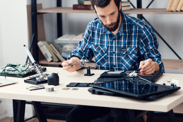 Electronics repairman workplace. Bearded engineer sitting at desk with special tools and fixing broken device