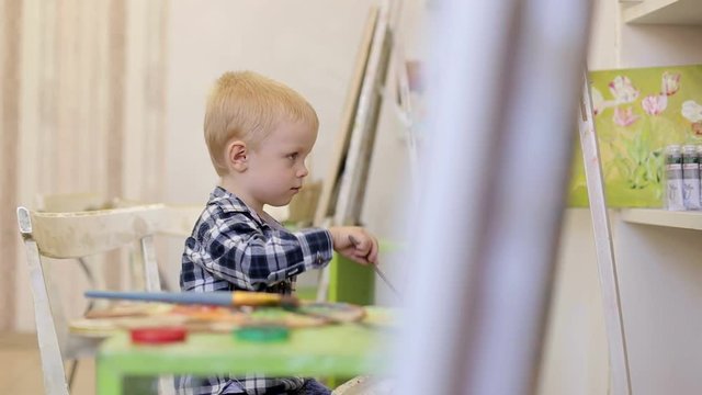 Cute boy painting a picture. Boy sitting on a chair in front of his easel in drawing school.
