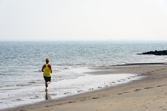Backlit image of a woman with blond hair pinned up, a yellow sport shirt, black shorts and sneakers running on the beach.