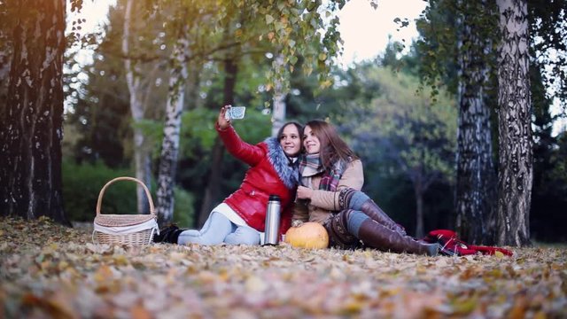 Beautiful girls making selfie on a picnic in autumn park sitting on the fallen leaves near the pumpkin at halloween time. 1920x1080