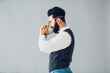 Young handsome bearded caucasian man posing indoors. Perfect skin and hairstyle. Wearing vest, white shirt, jeans. Studio portrait with dramatic light.