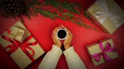 Top view hands drinking coffee christmas presents table on red background from above.