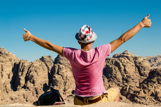 Self-satisfied man in hat on top mountain with thumbs up. Do not give up motivation concept. Happy hiker winning reaching life goal, success, freedom and achievement.