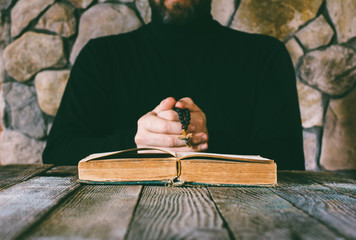 a man in black clothes with a prayer beads in hand praying in front of an old open book. the...