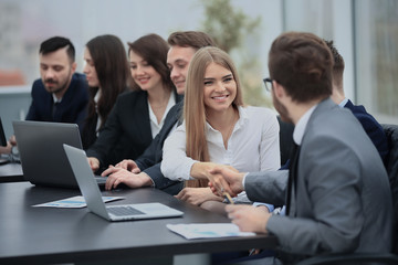 Group of confident business people in formalwear sitting at the table together and smiling while two men handshaking