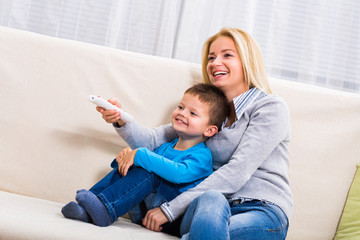 Happy mother and son are sitting on sofa and watching tv together.