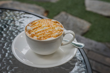 hot fresh coffee in white cup with caramel sauce foam on glass table in garden on grass and rock at coffee time