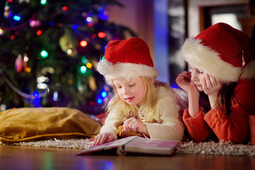 Fototapeta na wymiar Two cute little sisters reading a story book together under a Christmas tree