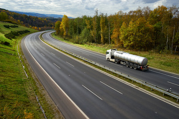 Asphalt highway with driving a silver tanker in the autumn landscape.