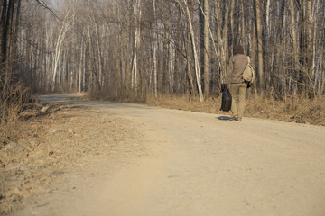 The hooded man with a backpack and a guitar goes on road near the forest