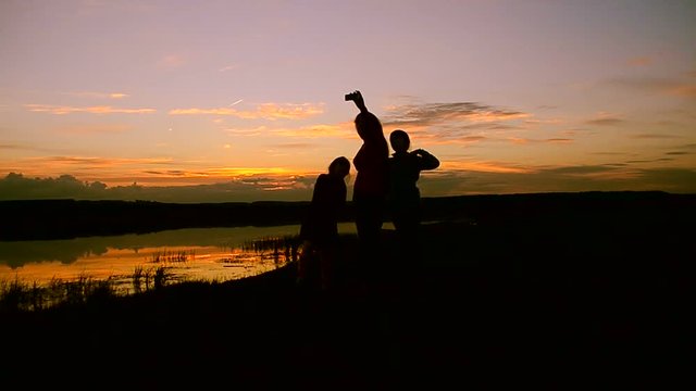 Young children with her mother on the phone taking pictures, make selfie on the banks of the river, lake, bay, against the backdrop of a beautiful sunset of the evening, the distant mountains, clouds