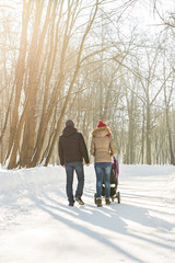 Happy young family walking in the park in winter. The parents carry the baby in a stroller through the snow.