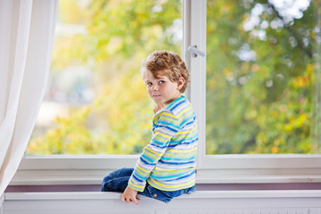 ittle kid boy sitting near window and looking on autumn trees