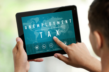 Young man using tablet. Text UNEMPLOYMENT TAX on screen. Online tax report concept.