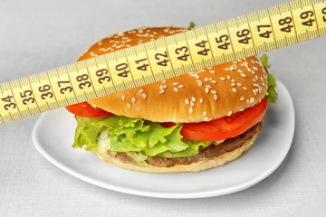 Dieting concept. Tasty hamburger on plate and measuring tape on light background