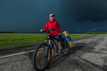 Fototapeta premium Lady hiker standing with loaded bicycle on the paved asphalt road with stormy clouds on the background