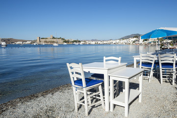 Fototapeta na wymiar Simple beachside tables line the pebble shore of the tourist resort of Bodrum, Turkey with a scenic view of the castle