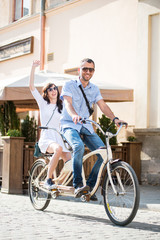 Fototapeta premium Young couple in sunglasses riding on tandem bicycle on the city at the sunny day. The man runs a bicycle, a girl waving hand