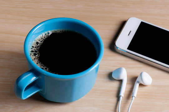blue cup of coffee and white earphones with smart phone on the wooden table