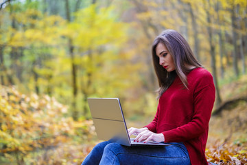 The beautiful woman sits in autumn park with the laptop