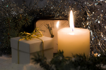 Christmas and New Year`s vintage clock showing five to midnight. White gift box with gold bow, festive evening candle, Christmas tree fir branches covered with snow. Background with holiday tinsel.