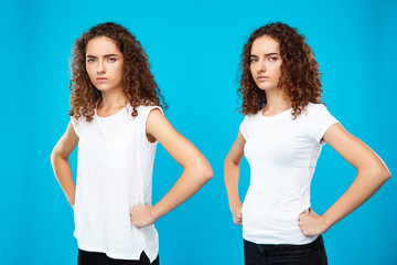 Two girls twins posing with arms akimbo over blue background.