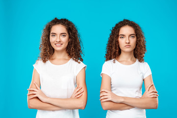 Two girls twins posing with crossed arms over blue background. - obrazy, fototapety, plakaty