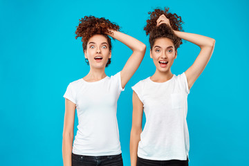 Two surprised girls twins holding hair, joking over blue background.