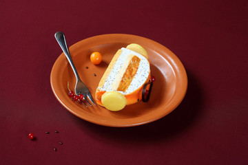 Piece of Contemporary Yule Log (layered) Cake covered with orange glaze and decorated with french macarons and chocolate loop, on brown plate, on burgundy background.
