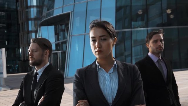 Slow motion. epic shot of three attractive confident businesspeople seriously look at camera. Sexy lady pomade brownhair front. Stand arm crossing modern building district bg. Teal and orange. Sunset.