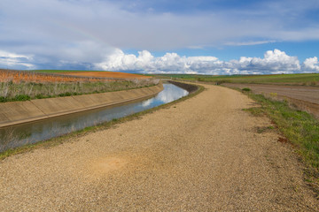 Fototapeta premium Canal de agua para el riego en curva y en paisaje agricola con camino asfaltado en paralelo 