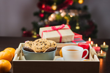Close-up of mandarins, candles, chocolate cookies, tea and gifts with Christmas tree on background