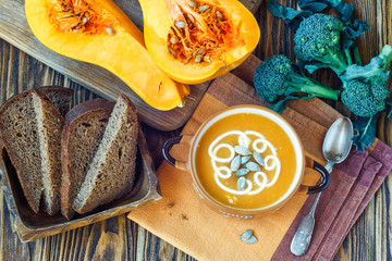 Roasted pumpkin soup with cream, fresh pumpkins and pumpkin seeds in plate on wooden background....