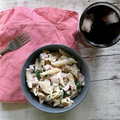Pasta penne is cooked al dente, then mixed with sliced ham, feta cheese and basil. Dressing is made with olive oil, bit of vinegar, salt and pepper. Served with glass of soda on white rustic table.
