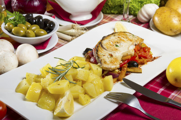 Potatoes side dish and fried fish steak on a white plate beside mushrooms, lemon and olives.