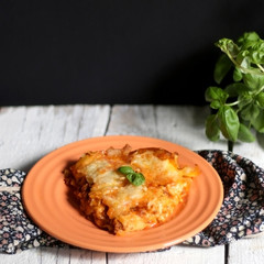 Homemade lasagna, made with Bolognese sauce and Bechamel sauce. Orange plate, white rustic table, decorated with some basil. Selective focus. 
