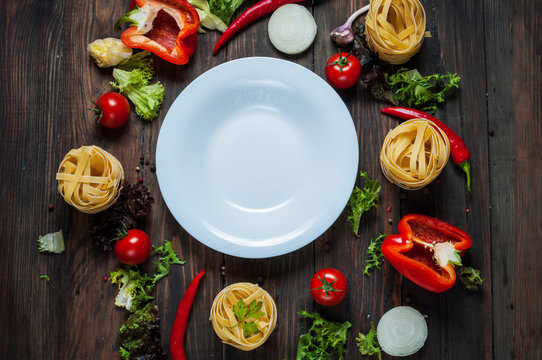 Ingredients for cooking spagetti, cherry tomatoes, pepper around a white plate place text, frame on wooden rustic background top view