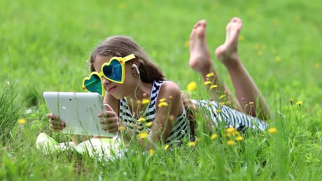 Pretty girl in big sunglasses with white tablet computer lies on green grass. Little girl in glasses lies on the green grass and uses tablet computer. Girl playing the game on his tablet computer

