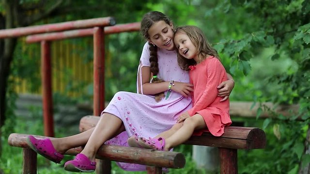 Two sisters sits on a wooden log at a sports ground and embraces. Girls sits together on a balance beam near parallel bars, communicates and embraces

