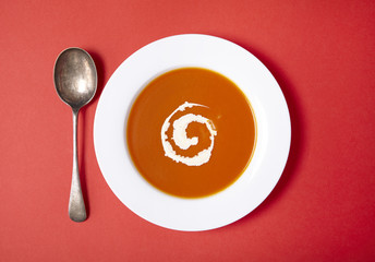 Aerial view of a bowl of hot tomato soup with swirl of cream and spoon on a bright red background