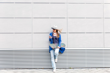 Young girl with skateboard leaning on modern gray wall, she is holding a mobile phone in her hand.