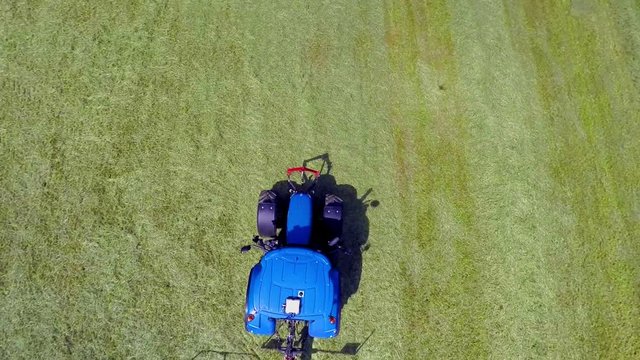 A big blue tractor has finished work and is now driving away from the fields. However, the rotary rakes are still moving. Wide-angle aerial shot.
