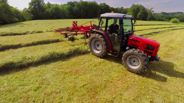 A red tractor which is pulling rotary rakes in the back is driving across lanes with hay. It is a nice summer day. Wide-angle shot.
