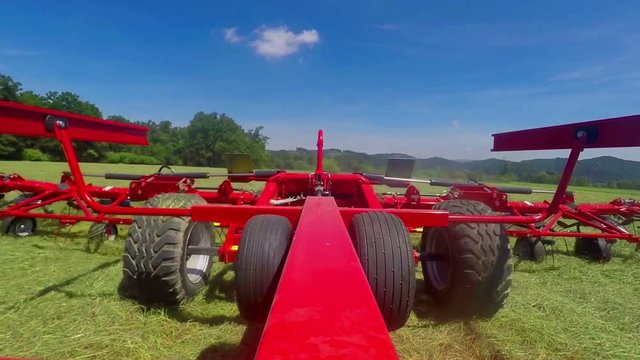A big tractor is pulling a huge machine with rotary rakes behind it. A farmer is working on a huge field and he is turning around a lot of dry grass. Wide-angle shot.
