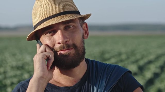 Tilt up of young bearded farmer with shovel standing on field and talking on mobile phone
