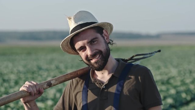 Medium shot of young bearded farmer with pitchfork posing for camera and smiling